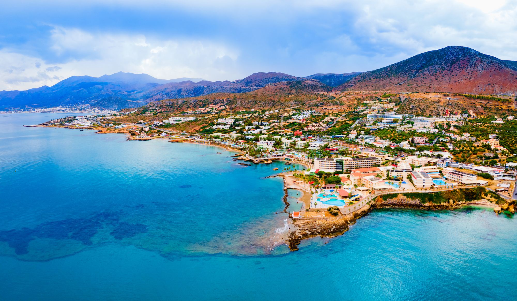 Bildet viser byen Hersonissos på Kreta sett ovenifra. Vi ser et blikkstille hav som beveger seg innover mot strandsonen av byen. Byen har spredt bebyggelse med grønne lunger som ender bakerst i bildet med fjellformasjoner.