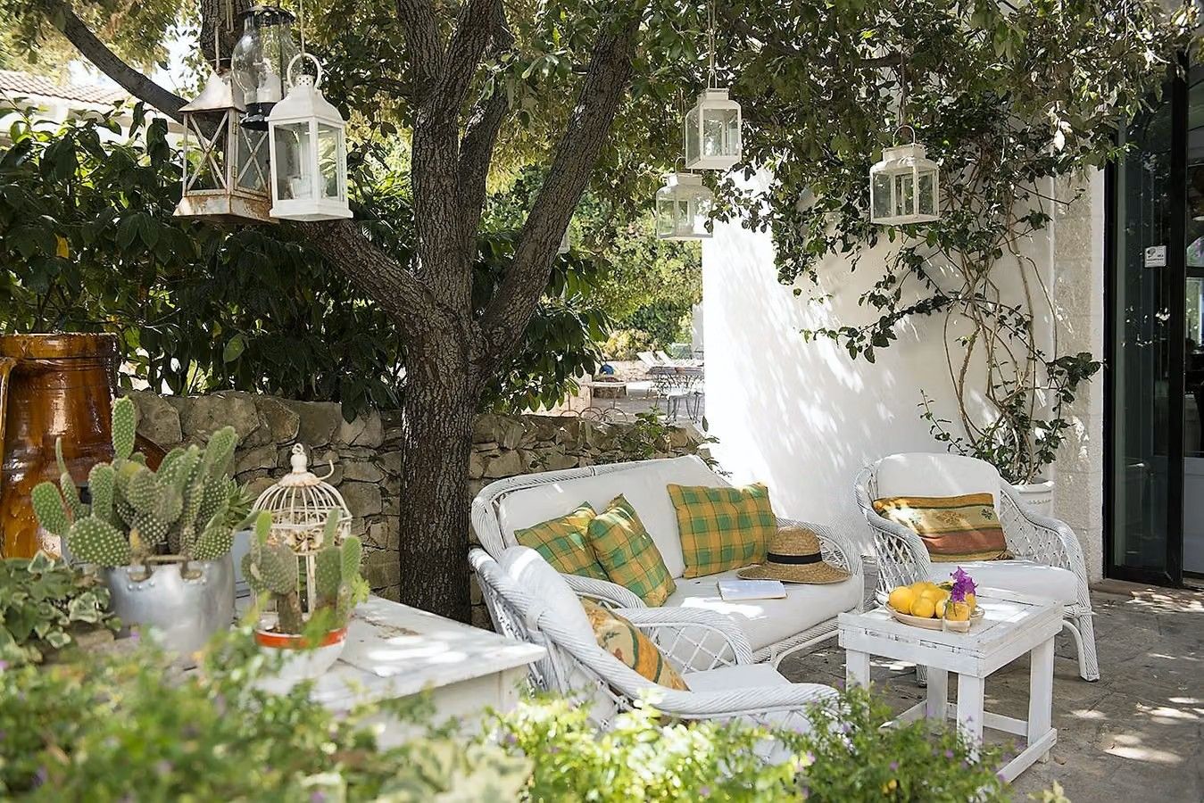 Bilden visar en del av utomhusområdet på hotellet Masseria Salinola i Ostuni i Apulien. Det är nästan som en privat trädgård, med vita trädgårdsmöbler, omgiven av träd, buskar, kaktusar och lyktor som hänger från träden.