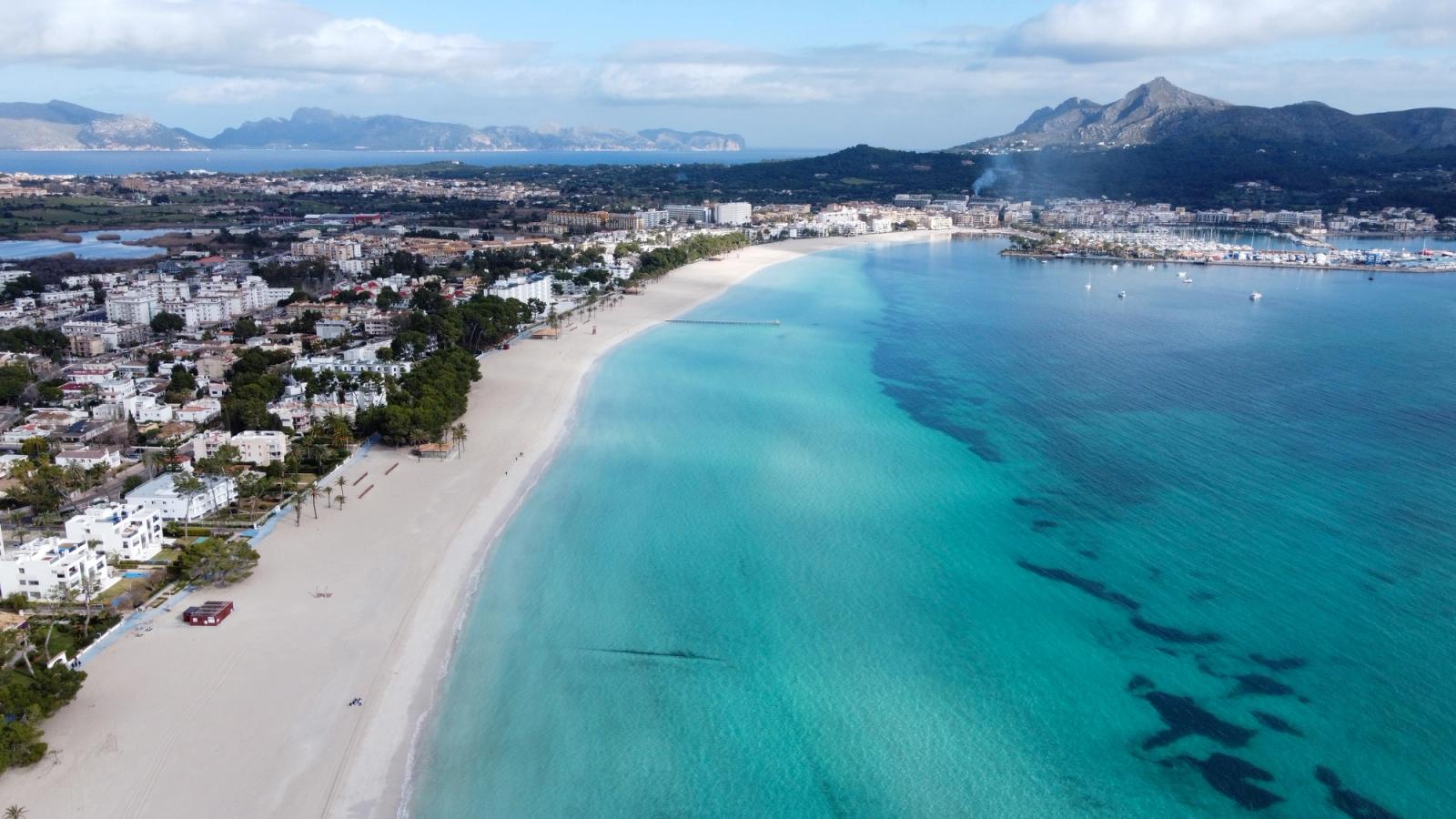 Bildet viser den langgrunne og langstrakte stranden Playa de Alcudia. Det blå krystallklare vannet til høyre og bebyggelsen bak stranden til venstre. Lengst inn i bildet kan man skimte bykjernen til Alcudia med fjelltopper bakenfor.