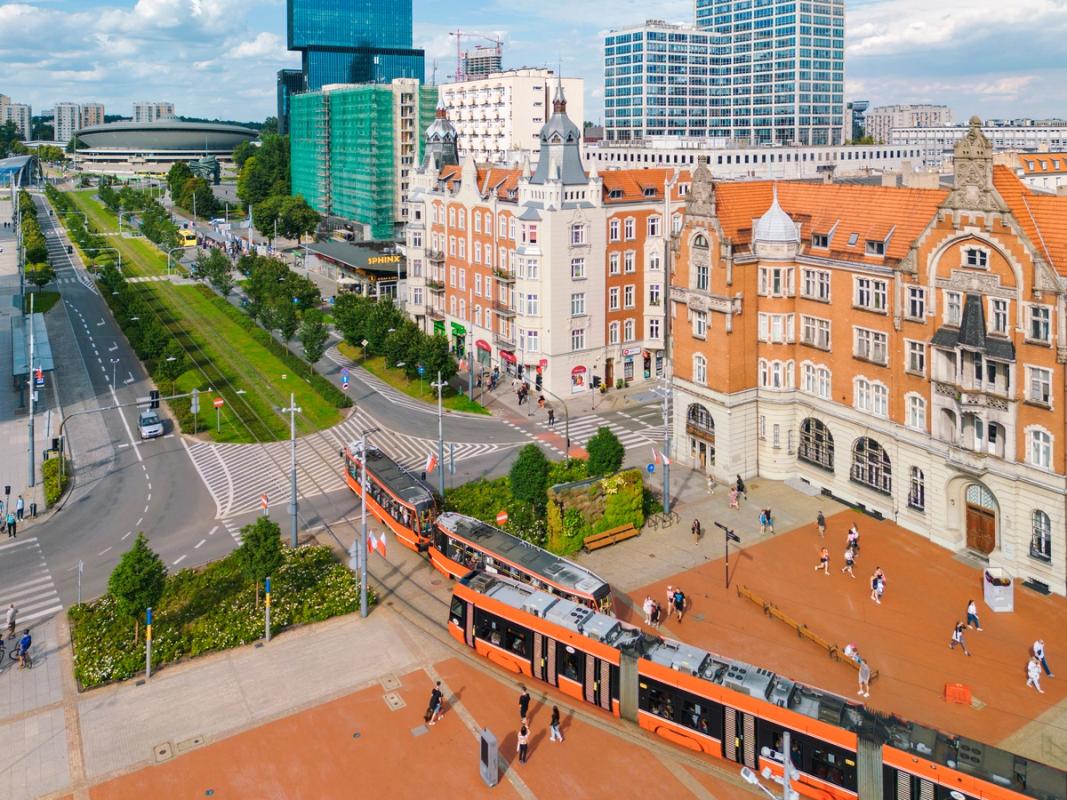 Bildet viser Katowice Rynek i sentrum av byen. Fremst i bildet vises to trikker på vei til og fra en grønnkledd aveny med trær på hver sin side. På høyre side eldre arkitektur som går over i en mer moderne arkitektur, med flere høyhus, lengre inn i bildet. På den åpne plassen vandrer mennesker på begge sider av trikkene.