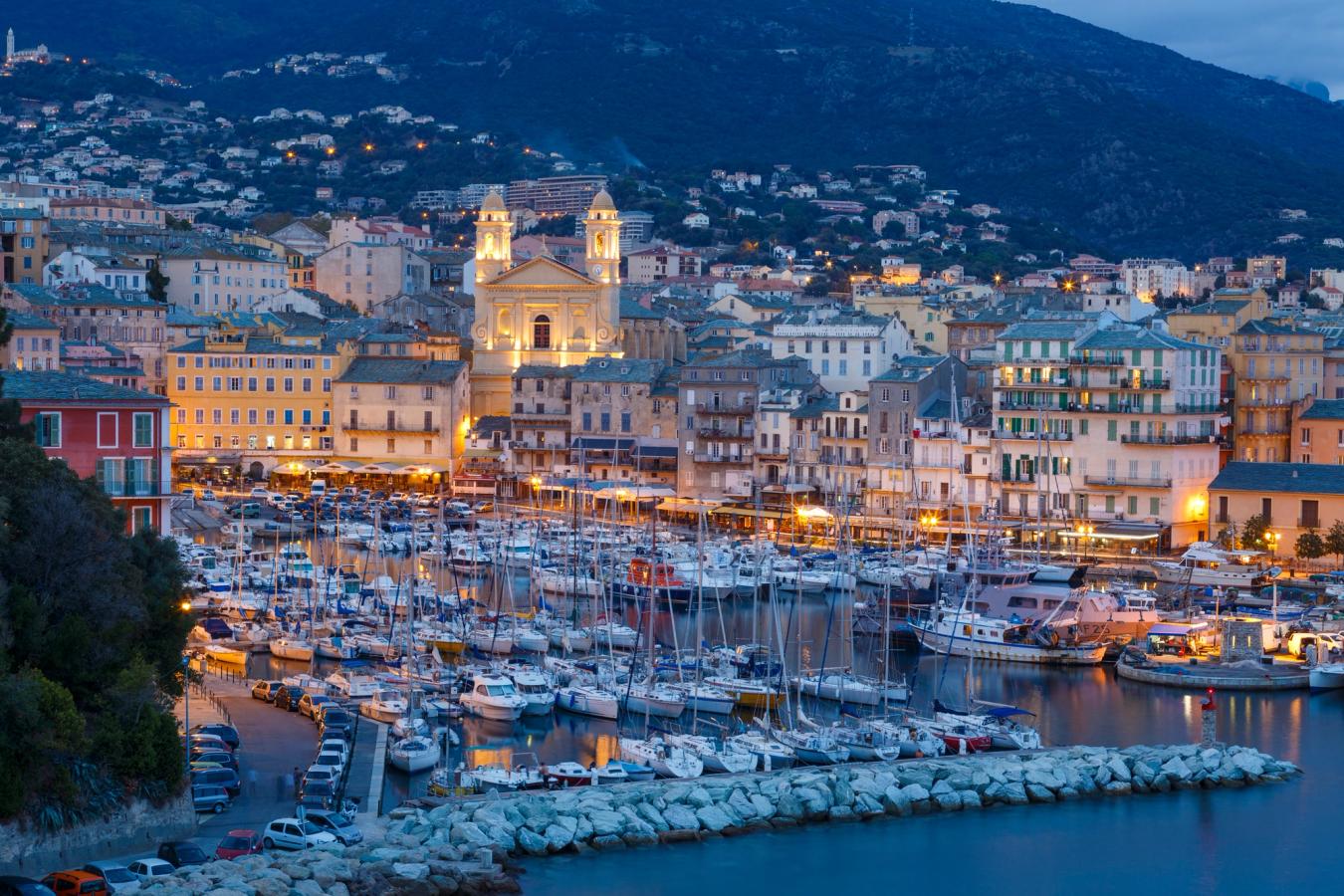 Bildet viser det gamle havneområdet i Bastia på kveldstid. Marinaen er fullt opp av båter og midt i bebyggelsen kan man se den opplyste St. John Baptist kirken, med sine to tårn.