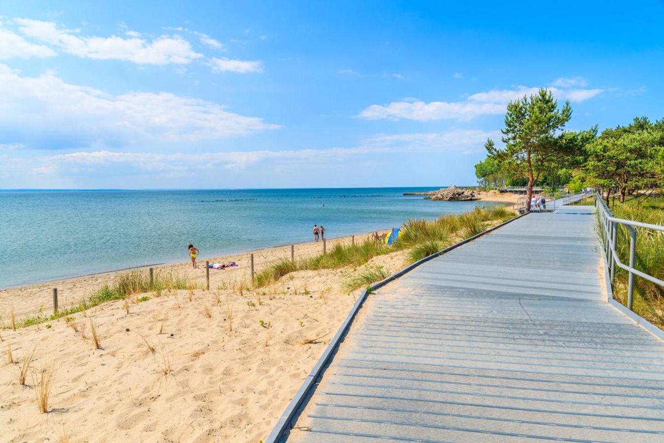 Langs en sandstrand med gyllen sand strekker det seg en trepromenade. På stranden er det få mennesker, og ved et furutre langs promenaden står noen sykler parkert. Havet er blått og strekker seg ut mot horisonten. Himmelen er blå med enkelte skyer.