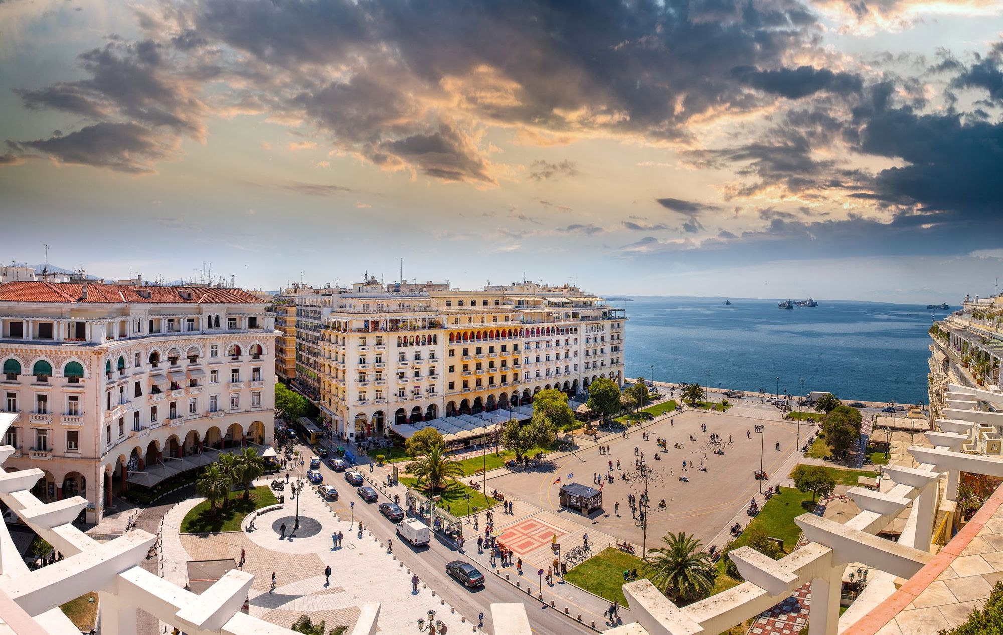 Bildet viser Aristotelous Square i Thessaloniki. En åpen plass med mennesker midt i. Plassen er først omgitt av grønn beplantning hvor plassen så blir omgitt av større bygninger, med bueganger på gateplan. Den åpne plassen ender ut i et stille hav.