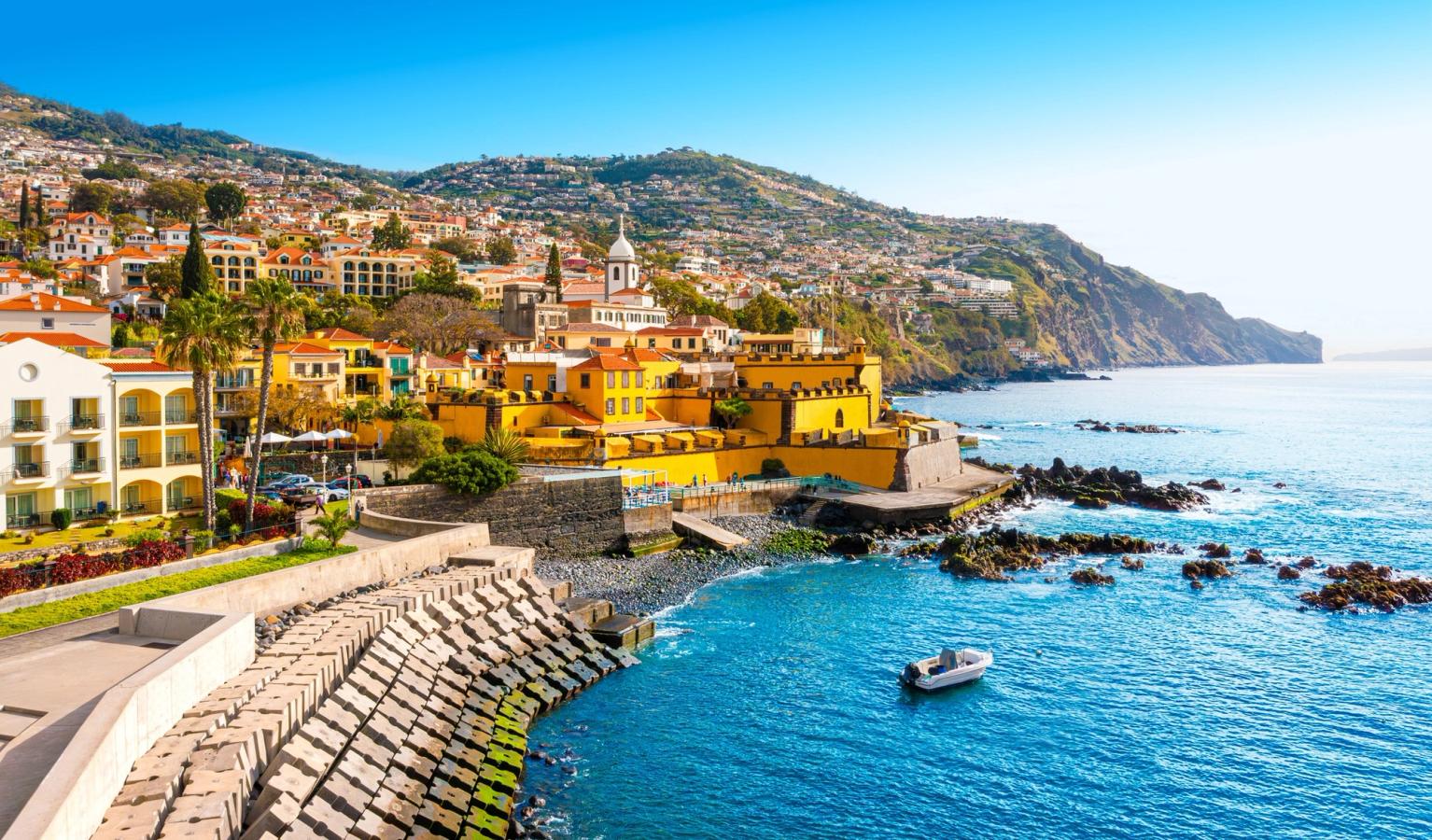 Bildet viser den gamle delen av Funchal, sett fra sjøsiden. Med lavere gulmalte hus oppover fjellveggen. Kystlinjen er av klippeformasjoner med blått hav.