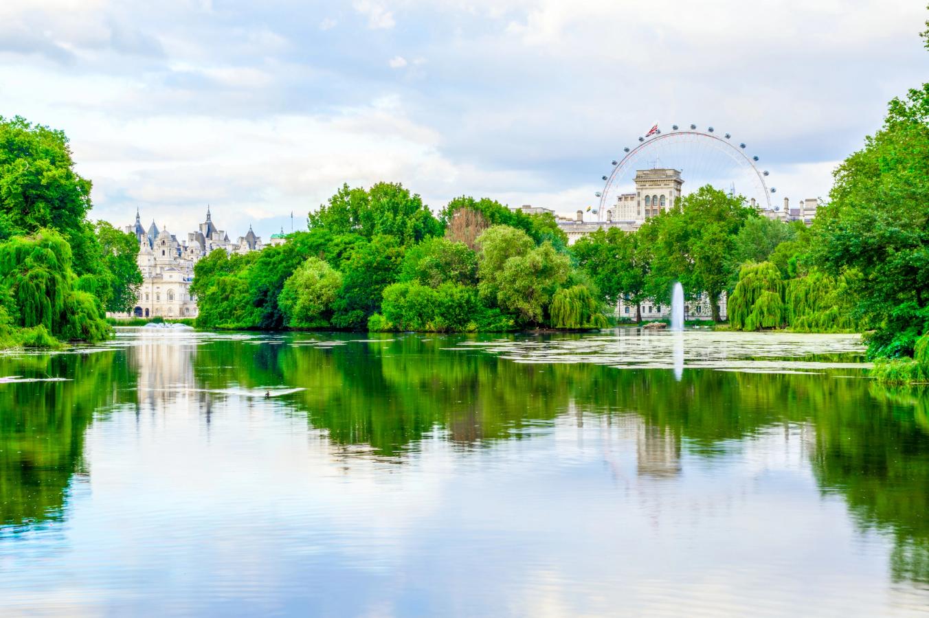 Bilden visar en vy från St James Park i London, där man ser ut över dammen i parken. Vattnet är omgivet av lummiga träd och olika näckrosor i vattnet. I slutet av bilden syns pariserhjulet London Eye till höger och den äldre arkitekturen kring Dover House till vänster