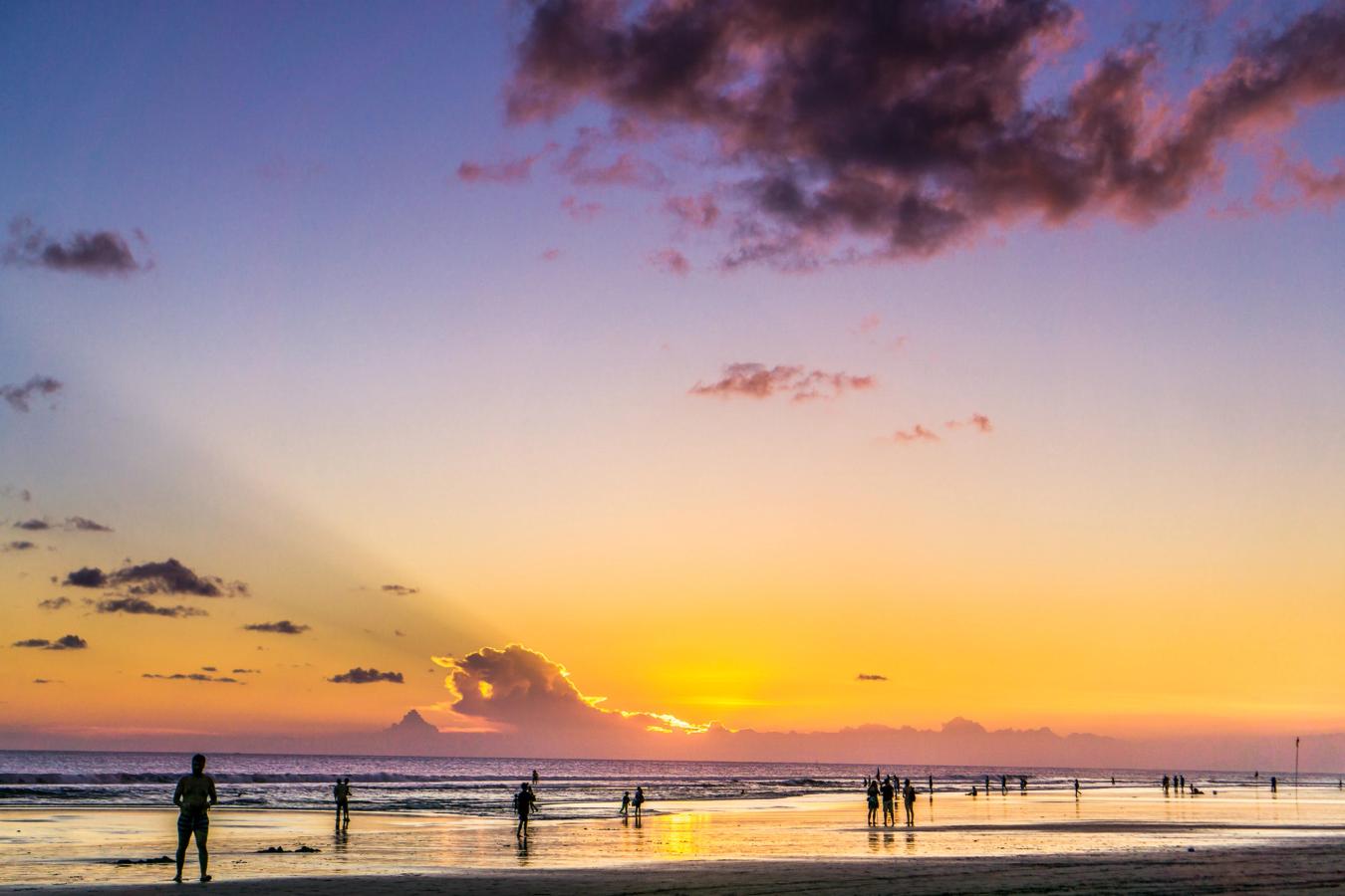 Flere mennesker går langs stranden imens solen er ved at gå ned. Himlen er oplyst i orange og lille nuancer. 