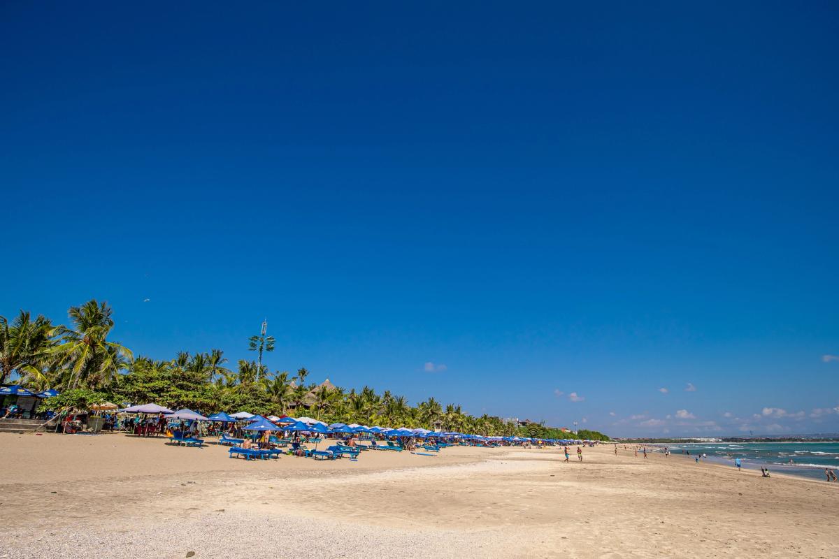 Gylden sandstrand i Kuta med blå parasoller og liggestole foran rækker af palmer. Flere mennesker går langs stranden, mens havet er turkisblåt under en klar blå himmel.