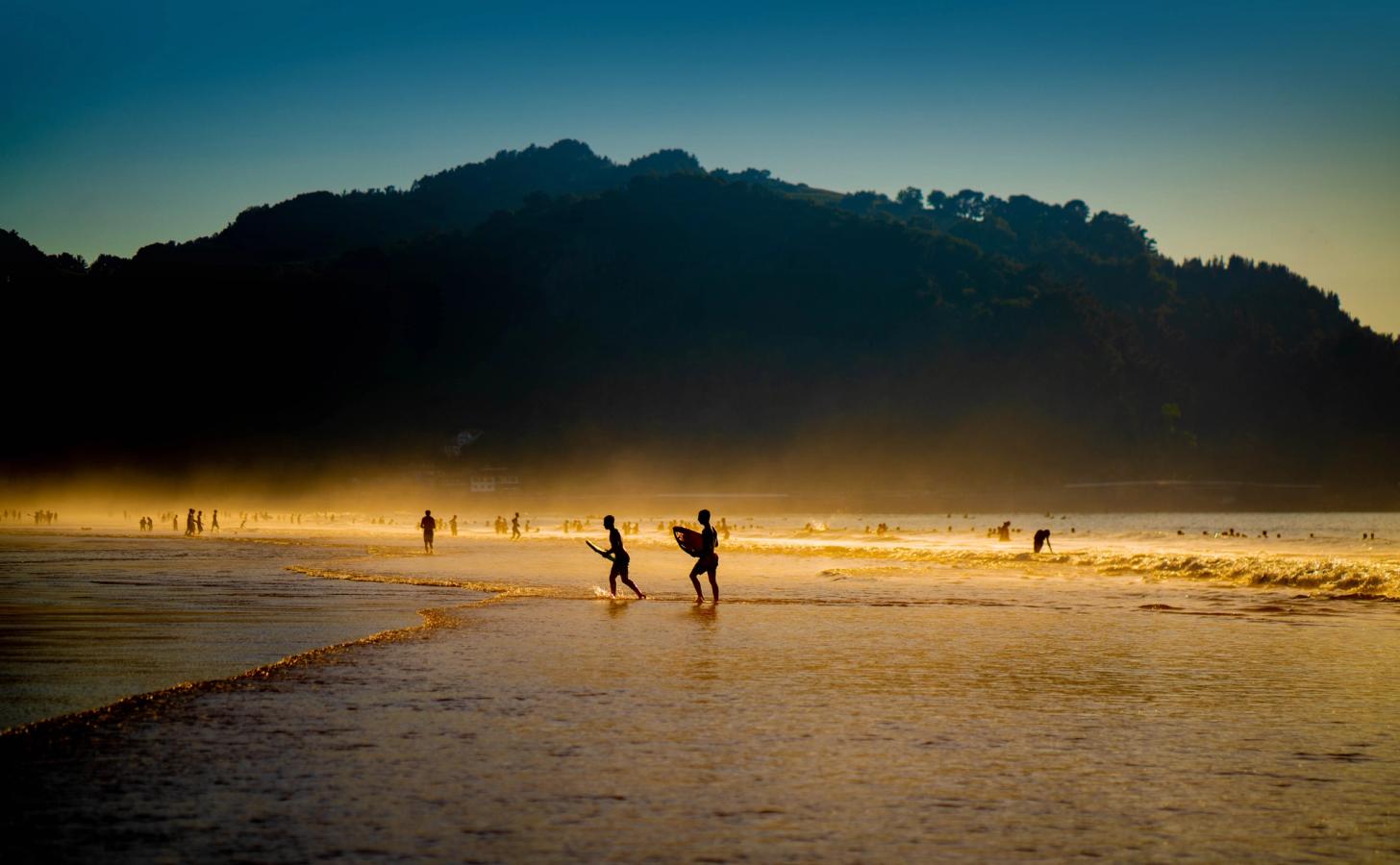 Stranden ved Zarautz i skumringstid. To personer med hver deres surfbræt går i forgrunden, mens flere mennesker opholder sig på stranden. Himlen er mørkeblå, og solnedgangen lyser sandet op.