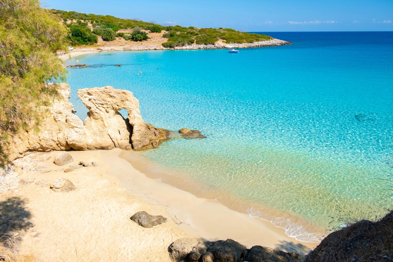 Utsikt over vakre Voulisma Beach, ikke langt fra Agios Nikolaos. Vi ser en lang sandstrand som deles i to av en klippeformasjon som går en anelse ut i vannet. Sjøen er stille med en klar blå farge. Bakenfor stranden er det en lavere og frodig natur.