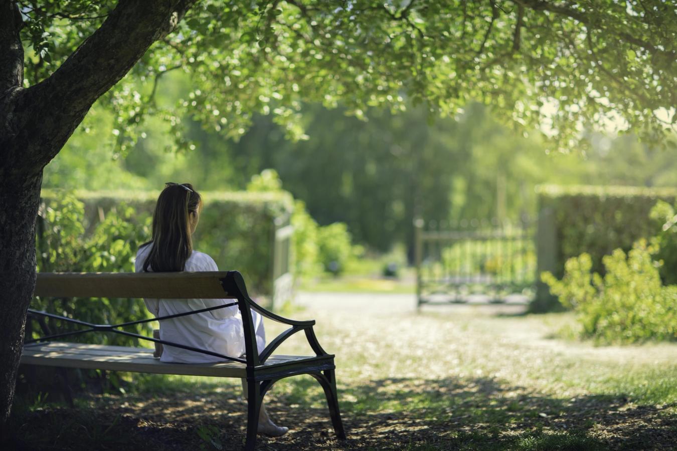 En ung kvinna sitter på en bänk under ett träd med ryggen mot kameran. Framför sig, i en lugn atmosfär med morgonsolen som lyser, ser hon ut över ett grönt parkområde