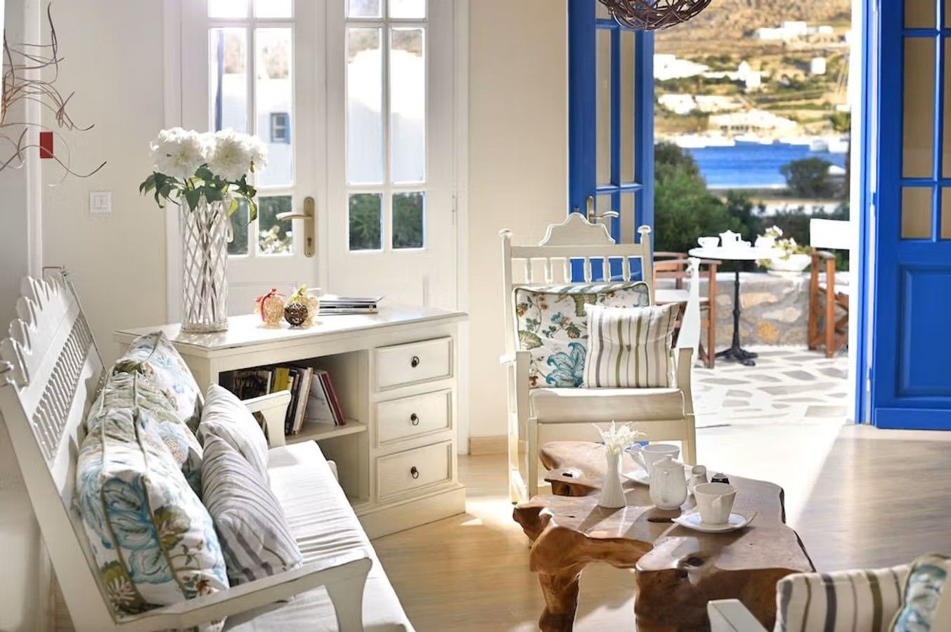 Innsiden av et rom på det familiedrevne hotellet Erato Hotel Mykonos. Møblene er hvite med bord og stoler, en terrassedør i koboltblå med videre utsikt over en terrasse og påfølgende sjø.