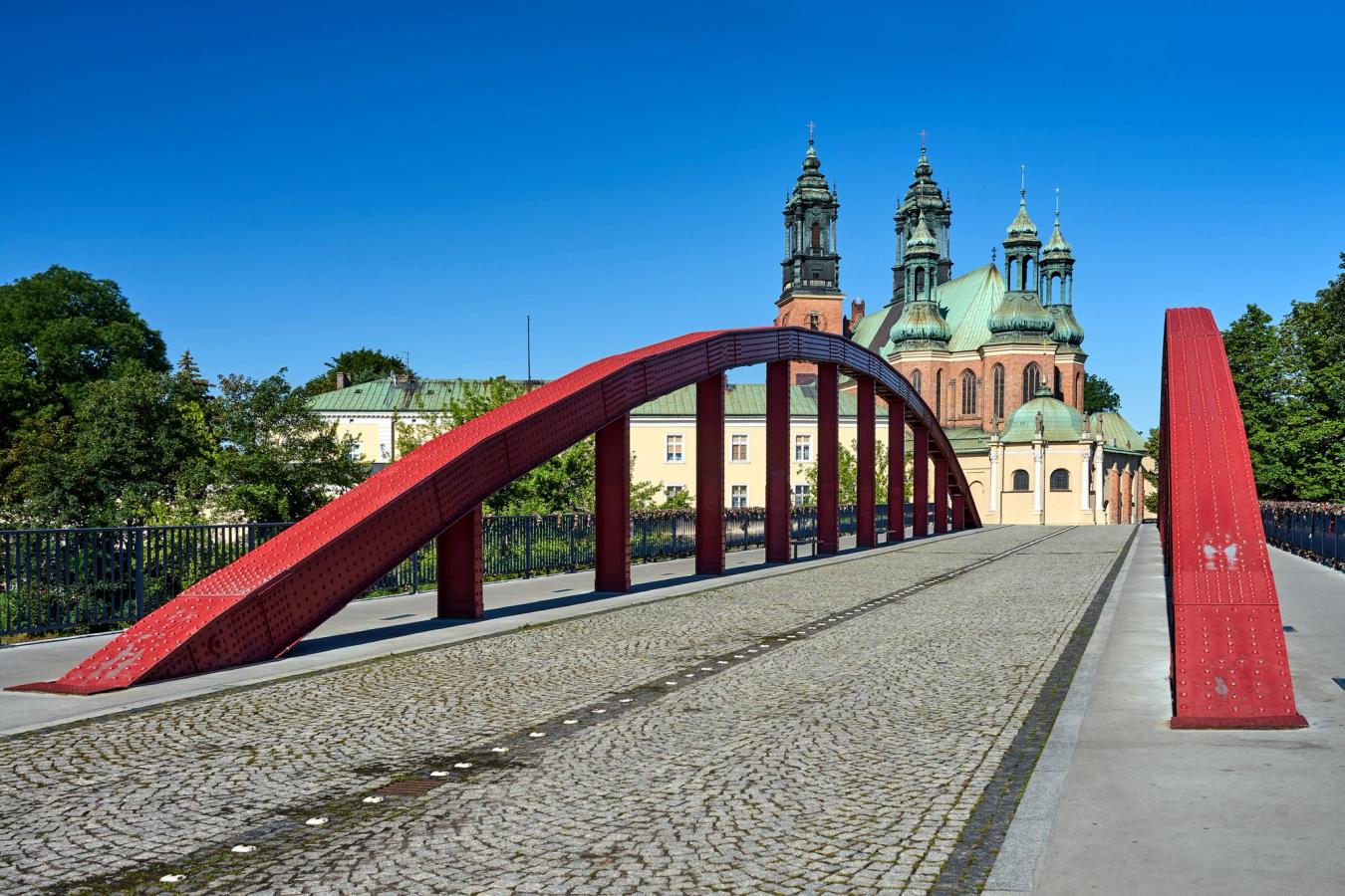 Fremst i bildet ser vi en brosteinsbelagt bro med rød smijerns arkitektur langs sidene. I enden av broen, lengst inn i bildet ser vi tårnene til Poznan Cathedral. Alt under en skyfri himmel.