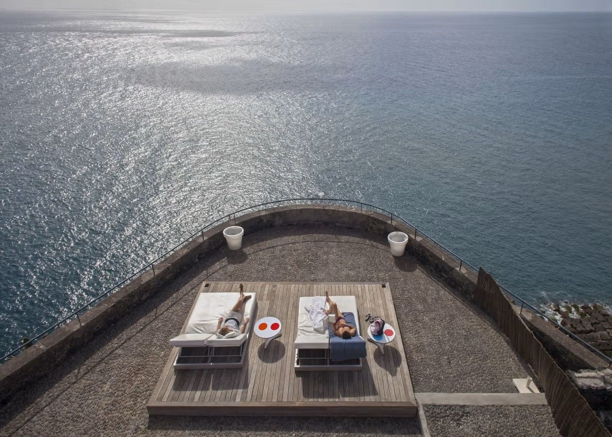 En større terrasse på hotellet Hotellet Estalagem Ponta do Sol på Madeira ligger på en klippetopp som gir en stor utsikt over Atlanterhavet. Midt på terrassen er det to mennesker på hver sin solseng som ser hele horisonten over det blå havet.