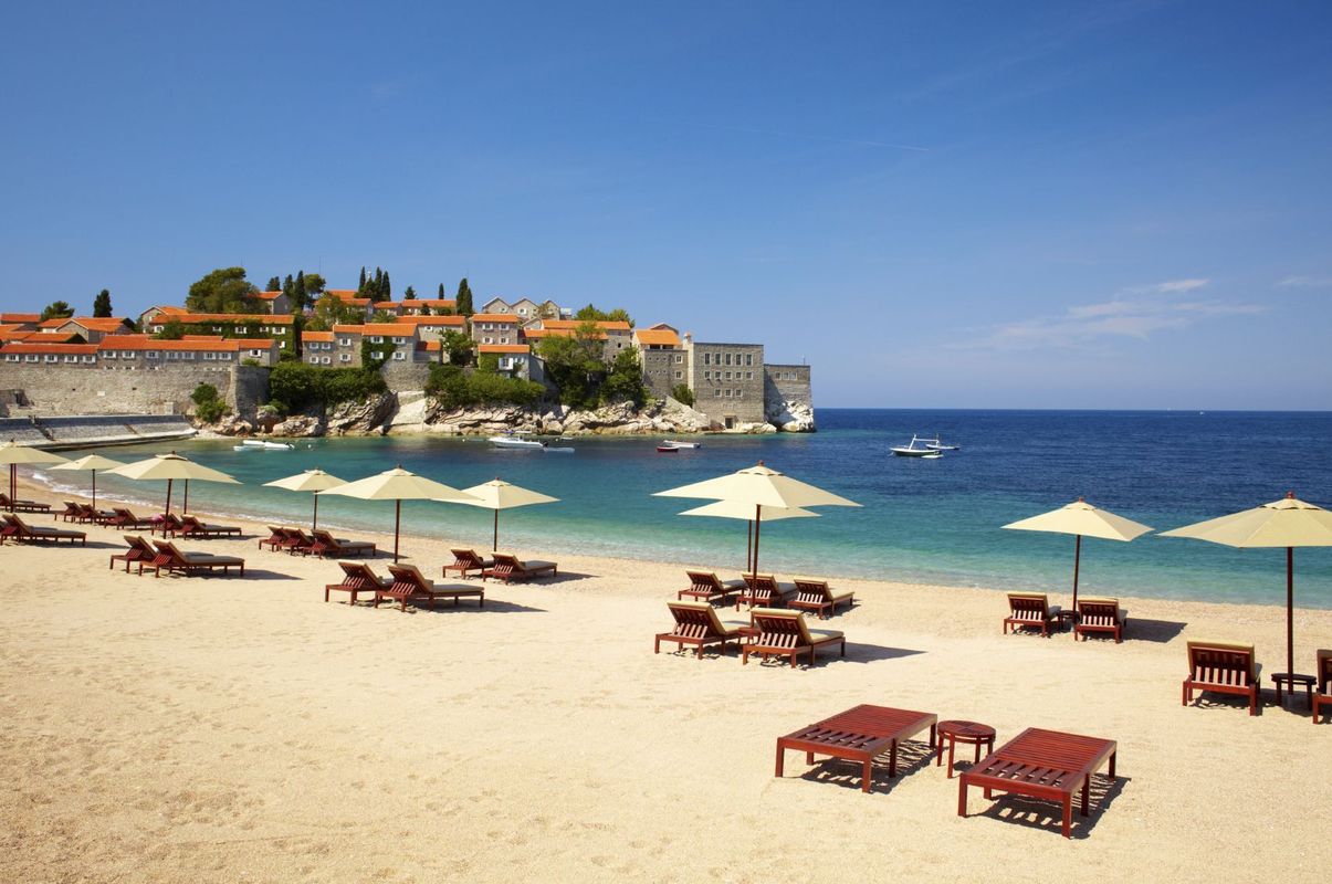 En strand med røde liggestole og hvide parasoller. Sandet er finkornet og vandet er turkisblåt. På vandet sejler en båd og i baggrunden ses gamle middelalderbygninger på øen ved Sveti Stefan.