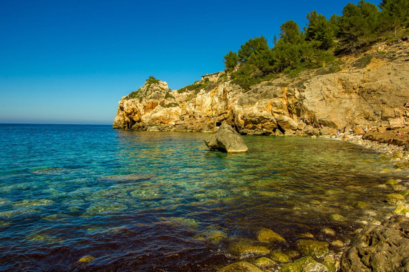 En lille stenstrand med krystalklart vand ved Puerto de Valldemossa. Stranden er omgivet af stejle klipper og himlen er blå.