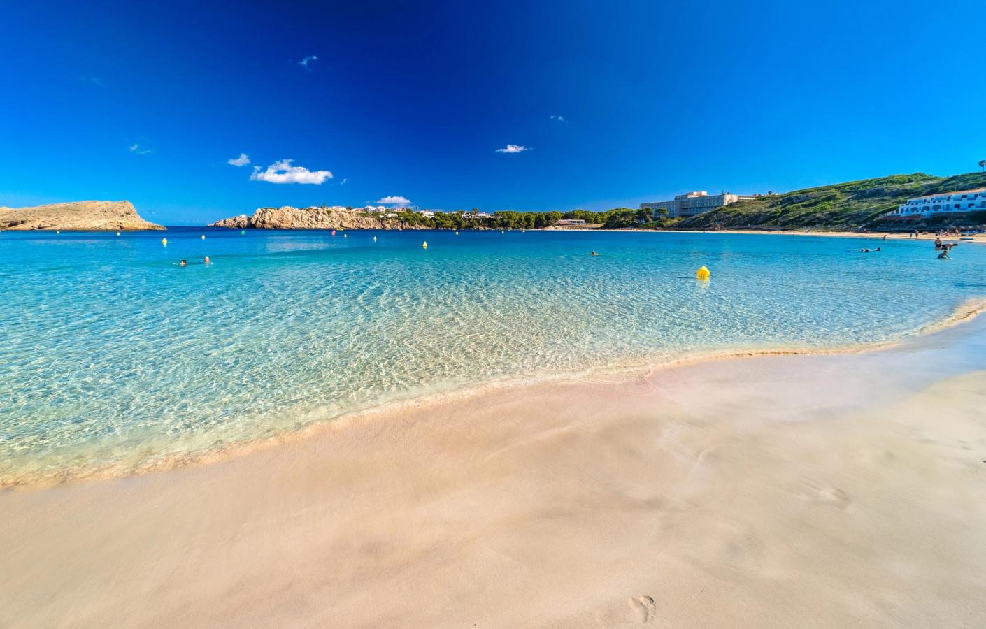 Billedet viser stranden i Arenal d’en Castell med hvidt sand i forgrunden og det klare vand i baggrunden. 