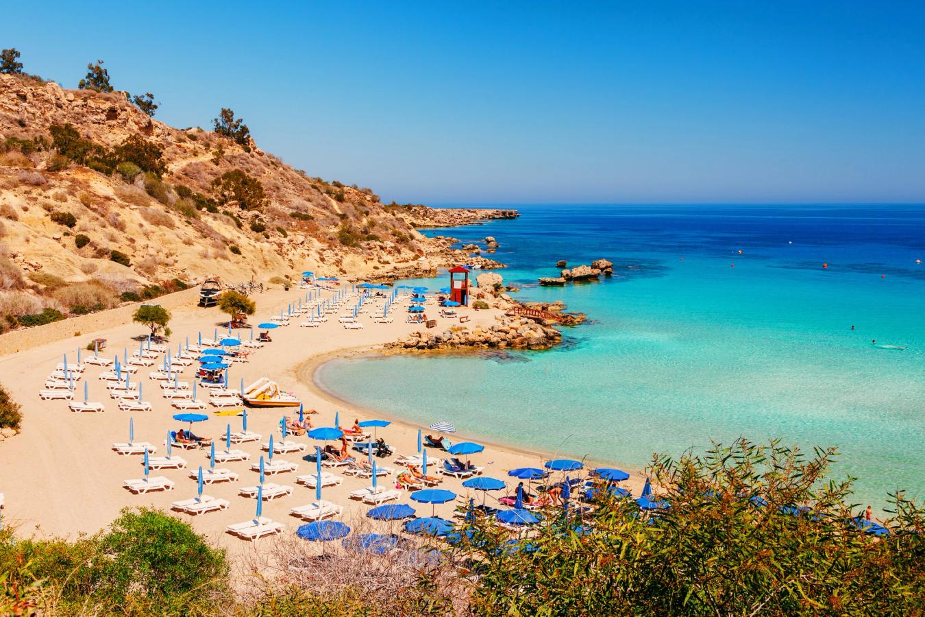 Bilden visar en mindre strand nära Nissi Beach på Cypern. Fin sandstrand med blå parasoller och solstolar. Längre till höger i bilden möter stranden det turkosa Medelhavet. Allt under en molnfri himmel