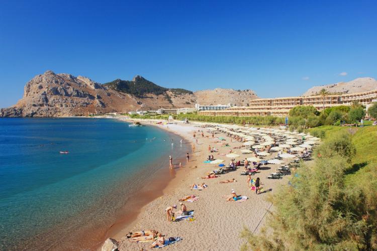 Bildet viser stranda i Kolymbia på Rhodos. Til høyre i bildet hotellbebyggelsen som går over i stranda med parasoller og mennesker som nyter sola. Badegjester i still vann til venstre under en skyfri himmel