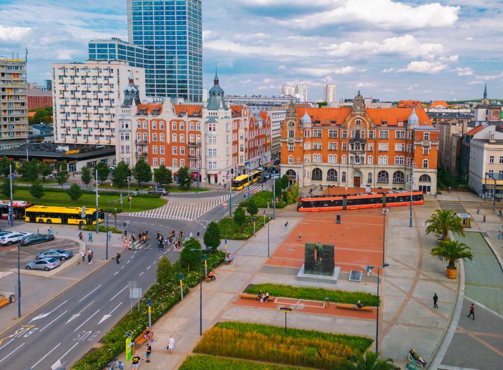 Billedet viser pladsen Katowice Rynek i centrum af byen. Forrest i billedet ses en bredere allé og gågade med beplantning. Gågaden munder ud i en plads med sporvogne og anden kollektiv trafik samt ældre bebyggelse og arkitektur. I baggrunden ses flere højhuse og et tættere bycentrum. Spredte skyer på himlen.