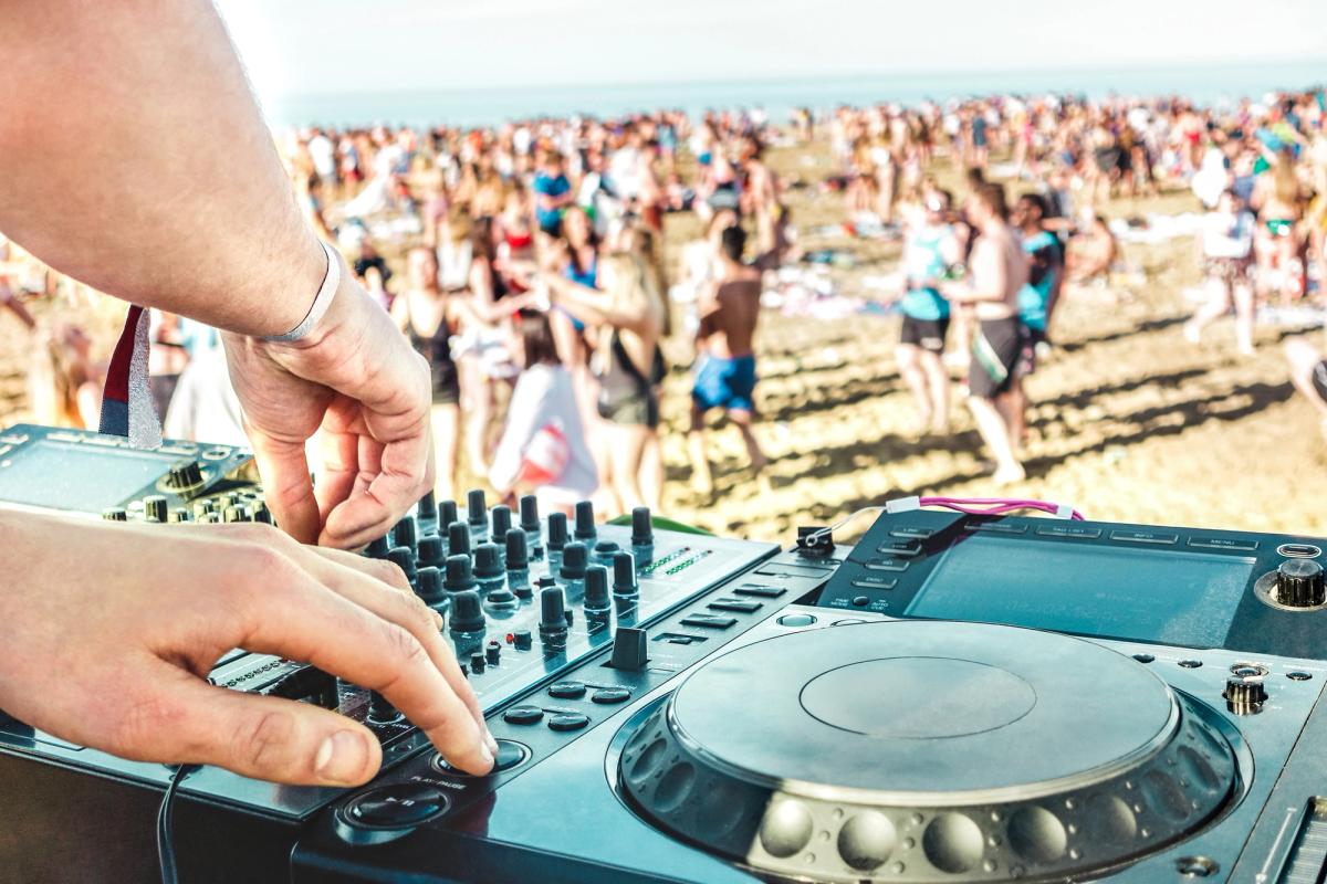 Bilden visar två händer från en DJ som arbetar över ett mixerbord. I bakgrunden många festglada människor på stranden på dagtid.