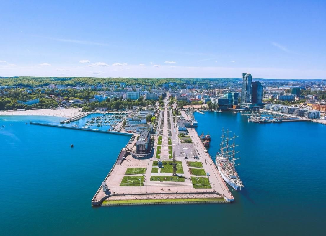 Bilden visar i mitten den populära piren, eller brygga, i Gdynia som sträcker sig långt ut i havet. Piren har en blandning av parkdekorationer och promenadstråk längs sidorna. Till vänster ser vi stadsstranden och längre in i bilden mer av själva bebyggelsen i Gdynia.