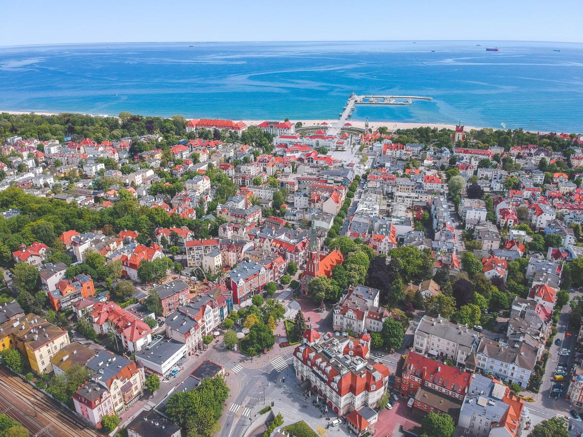 Bildet viser et oversiktsbilde av Sopot by. Her vises byens bygningsmasse med klassiske røde tak og spredt grønn beplantning i gatene. Byen ender ut i sandstranden langs den blåe Østersjøen.