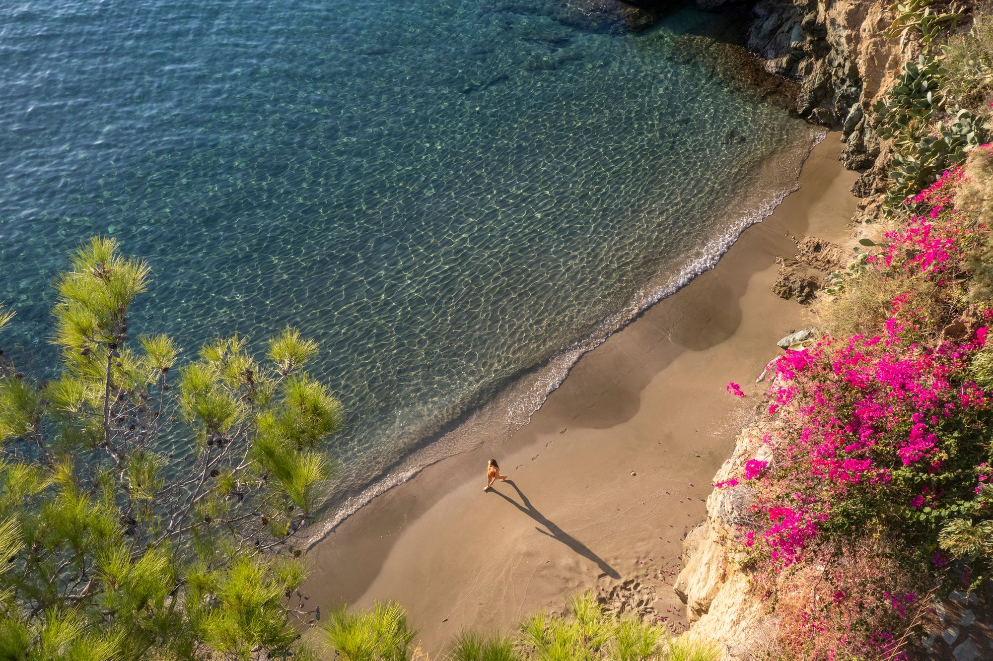 Bildet viser en liten og smal strand ved Agia Pelagia på Kreta. Bildet er tatt fra klippehøyden over stranda og langs vannet kan man se en kvinne spasere. Nedover klippeveggen til høyre i bildet ser man vakre røde blomster som gror ned fra fjellet. Stranden er svært smal med rolig vann som bølger svakt over sanden