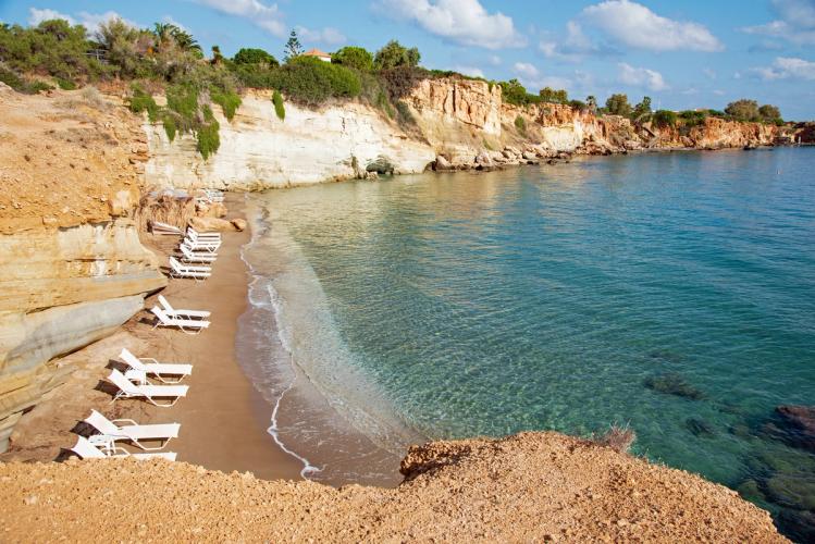 Utsikt over en del av stranda Sarantari Beach ved Anissaras på Kreta. Den lille biten av stranda er avskilt fra resten med klipper ut i sjøen. Selve stranda er smal med utplasserte hvite solsenger. Bak og omkring stranda, i høyden ser man grønn vegetasjon. Stranda er langgrunn med et grønnaktig skjør i det stille vannet.