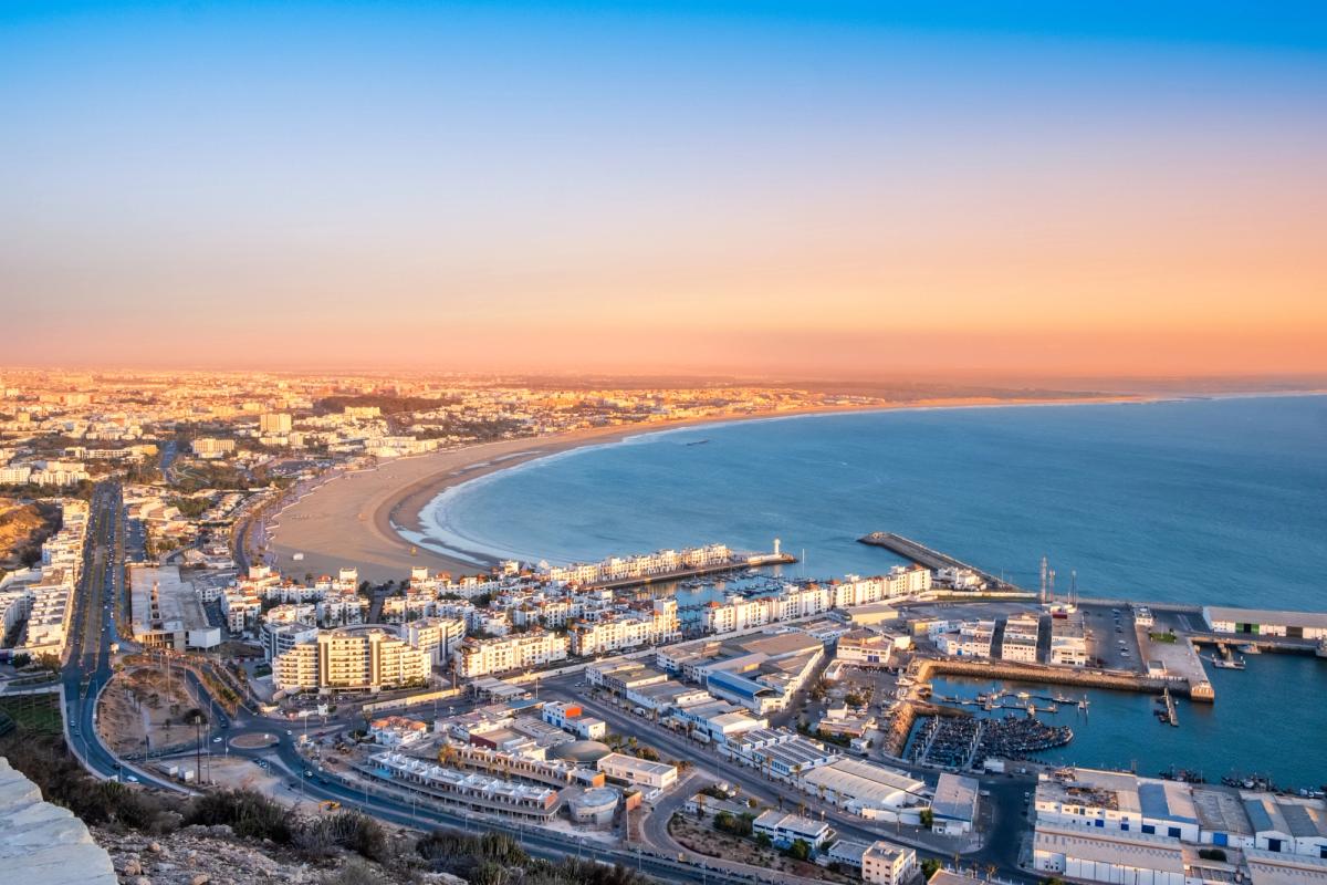 Drönarbild över stranden och bukten utanför staden Agadir. Bakom stranden ligger tät bebyggelse under en molnfri himmel med kvällssol