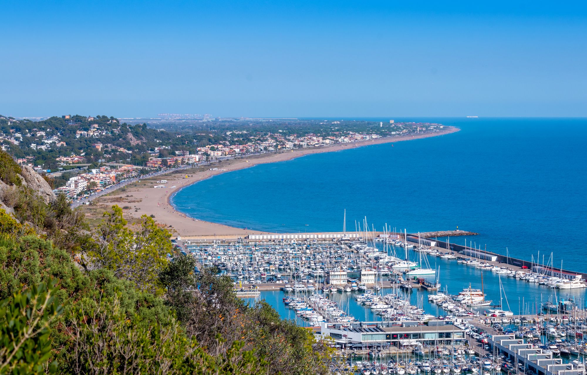 Utsikt över marinan i Castelldefels. I förgrunden löper grön vegetation längs en sluttning, och vid horisonten smälter himmel och hav samman till ett.
