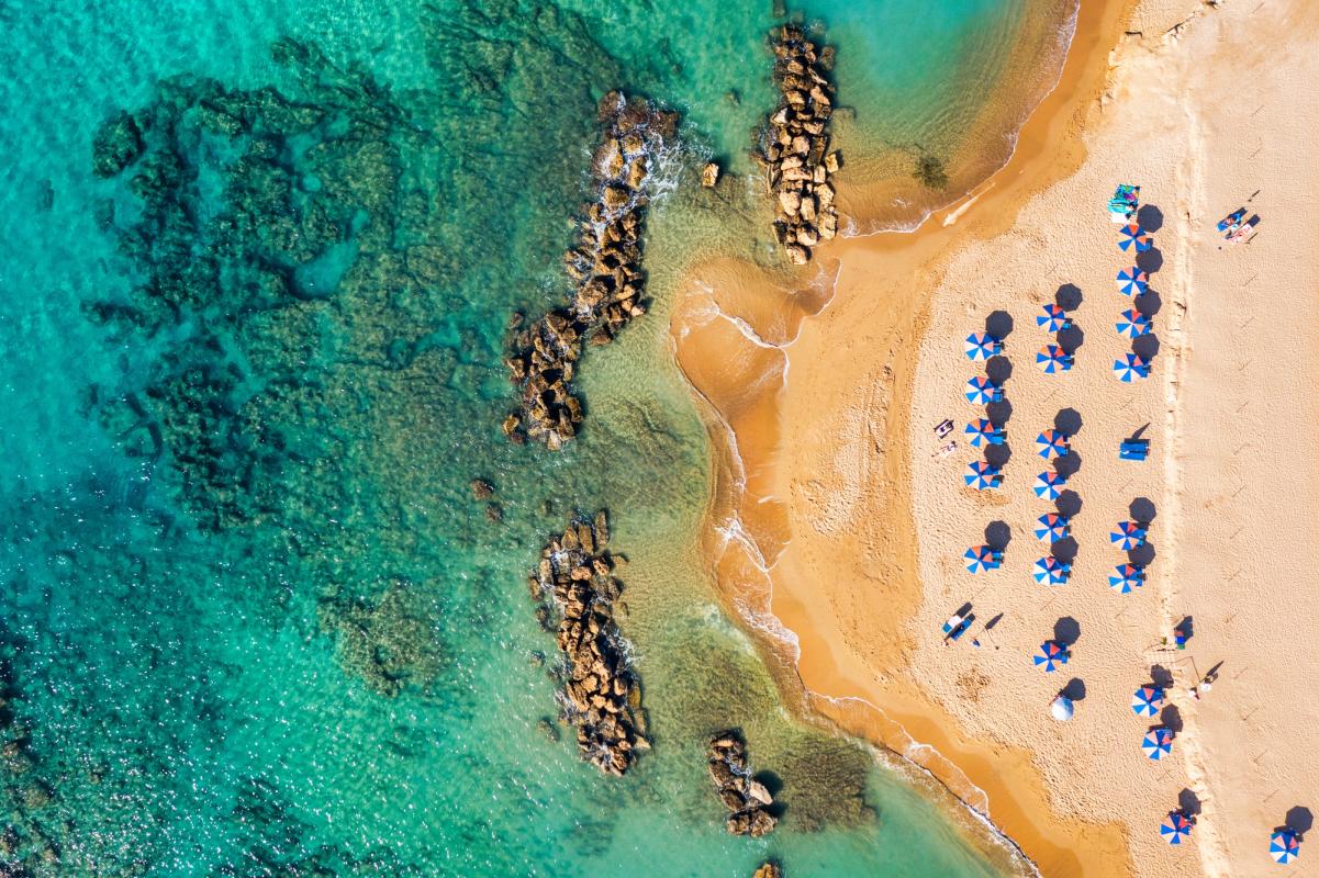 En gyllene sandstrand med rader av blå parasoller och solstolar. Till vänster syns det turkosblå havet med mörka klippformationer under vattnet och längs kusten.