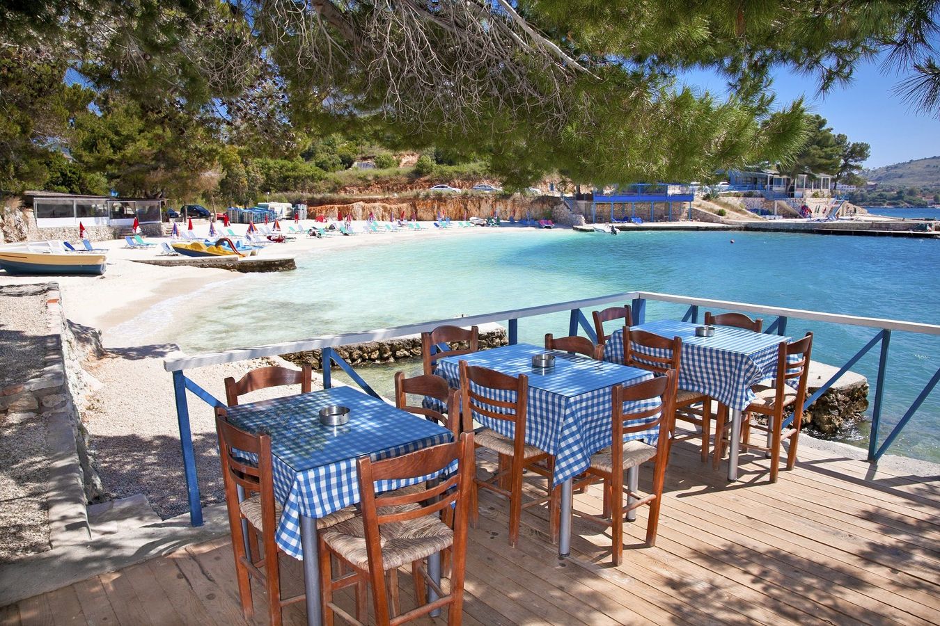 Bildet viser tre bord med blårutede duker på en kafe i Ksamil. Ved siden av kafeen strekker stranden seg innover i bildet med stille blått vann.