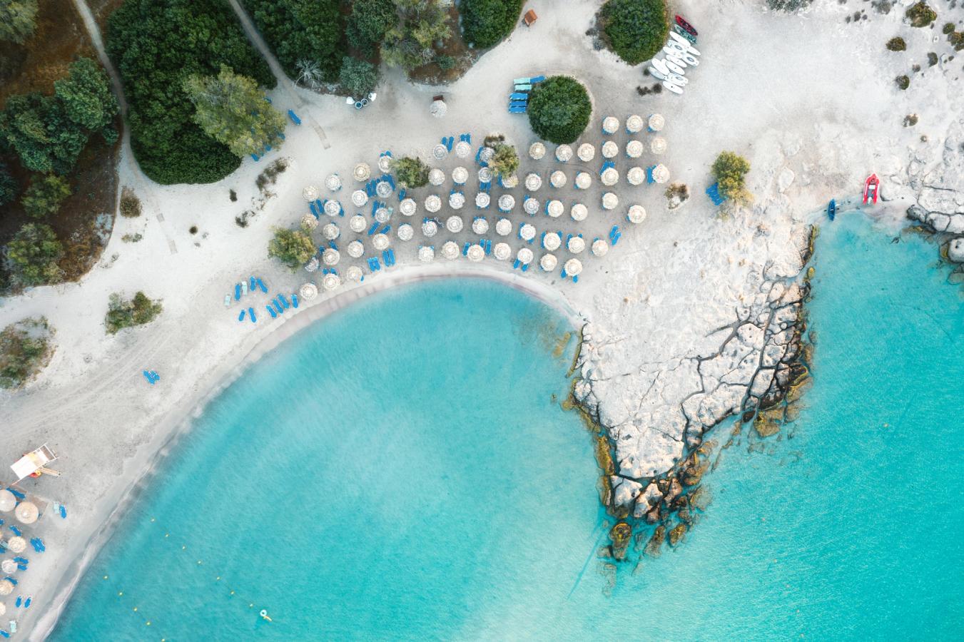 Bilden visar Elafonissi-lagunen på den västra sidan av ön Kreta. Stranden är kritvit och slutar i kristallklart blått vatten. Vita parasoller är uppställda på stranden och bakom stranden ligger en lummig liten skog