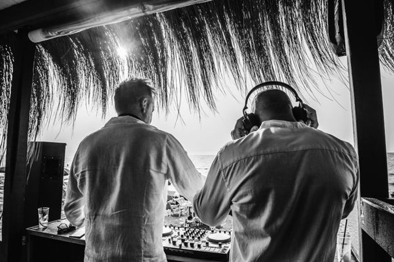 Bilden visar ryggen på två DJ:s som spelar på en strand. De står i skuggan under palmtak med mixerbord framför sig. Bilden är i svart/vitt