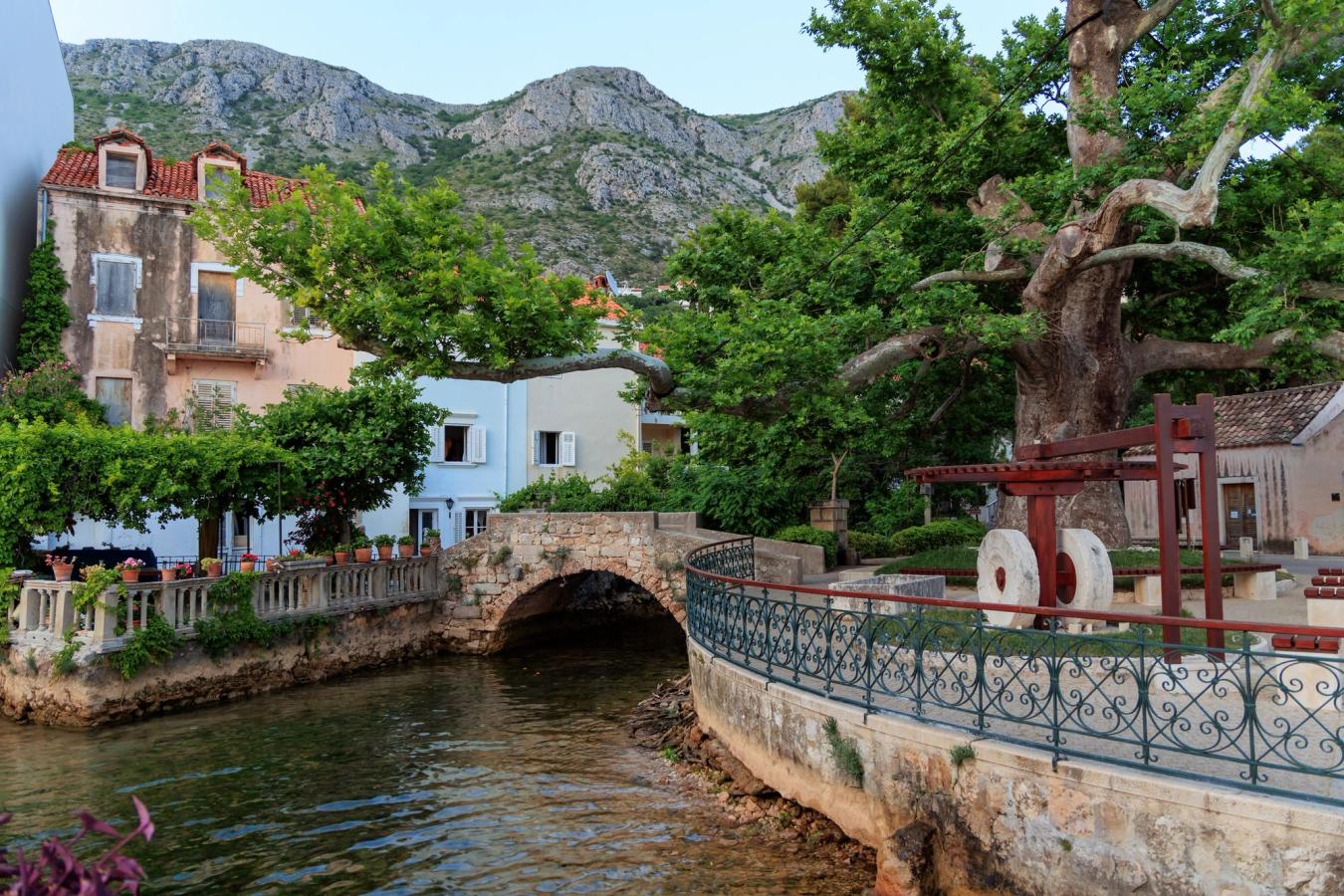 En liten kanal renner gjennom byen. På høyre side står en gammel vannmølle, og til venstre er det hus i forskjellige farger. I bakgrunnen reiser fjellene seg.
