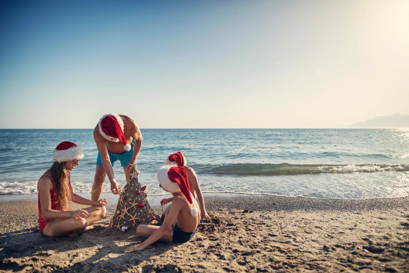 Fire personer sitter på en strand med nisseluer og lager et juletre av sanden