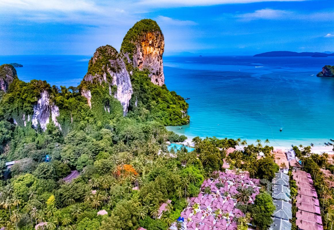 Bildet viser Railay Beach på Krabi, sett litt fra avstand. Nærmest i bildet ser vi en frodig skog med en rekke bungalower. Til venstre to høye klipper som ender opp i blikkstille blått hav.