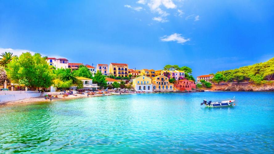 turkisblått hav foran en liten by med fargerike hus og frodig buskvekst i og ved siden av byen. Foran byen er det en smal strand med mange som soler seg.
