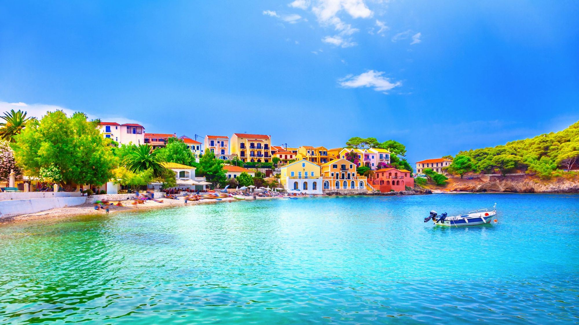 Turkosblått hav framför en liten stad med färgglada hus och frodig buskvegetation i och runt staden. Framför staden finns en smal strand med många som solar sig.