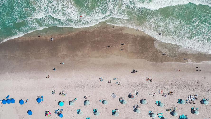 Parasoller og solsenger står på den gyldne sandstranden ved kysten, mens det blå havet glitrer i solen og bølgene ruller stille inn mot land
