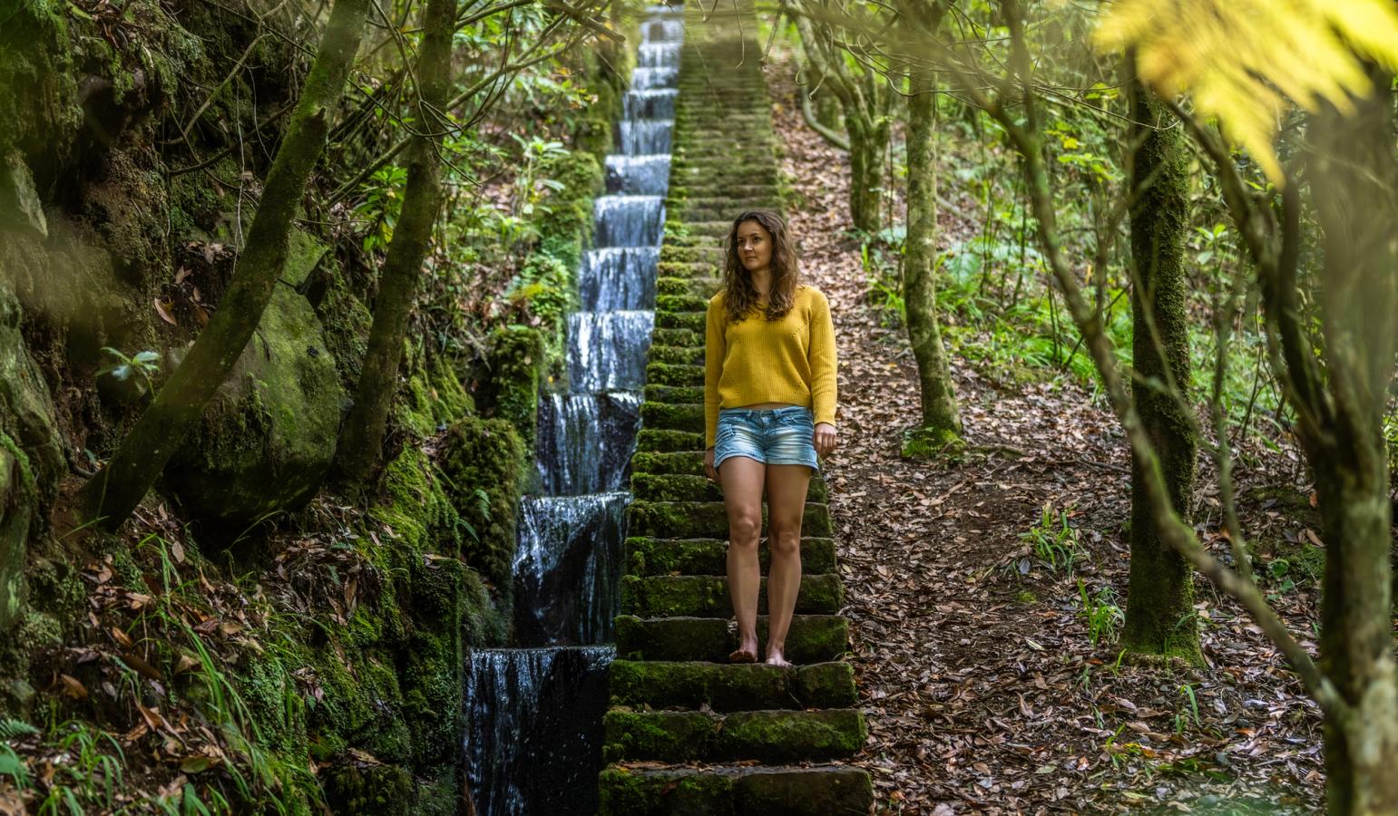 En kvinne som går nedover trapper langs Levada-stiene på Madeira. Vann renner nedover på hennes høyre side og på trappetrinnene gror det grønn mose. Kvinnen er ellers omgitt av lys og frodig skog.