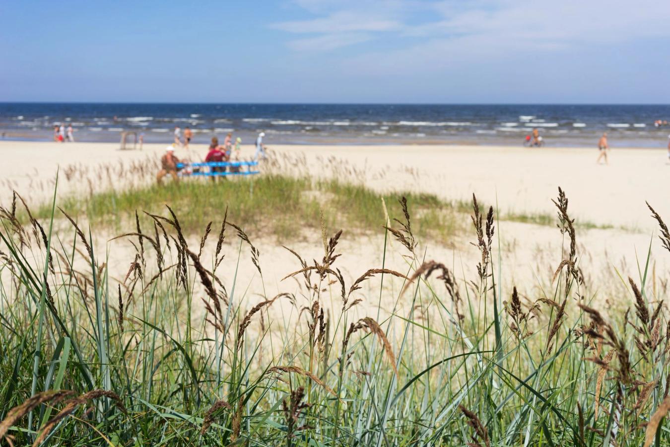 Sandstrand med siv i forgrunnen. Flere mennesker går langs vannkanten, mens et par sitter på en blå benk. Havet strekker seg mot horisonten med hvite bølger som ruller inn mot stranden. Himmelen er blå med et lavt skydekke. 