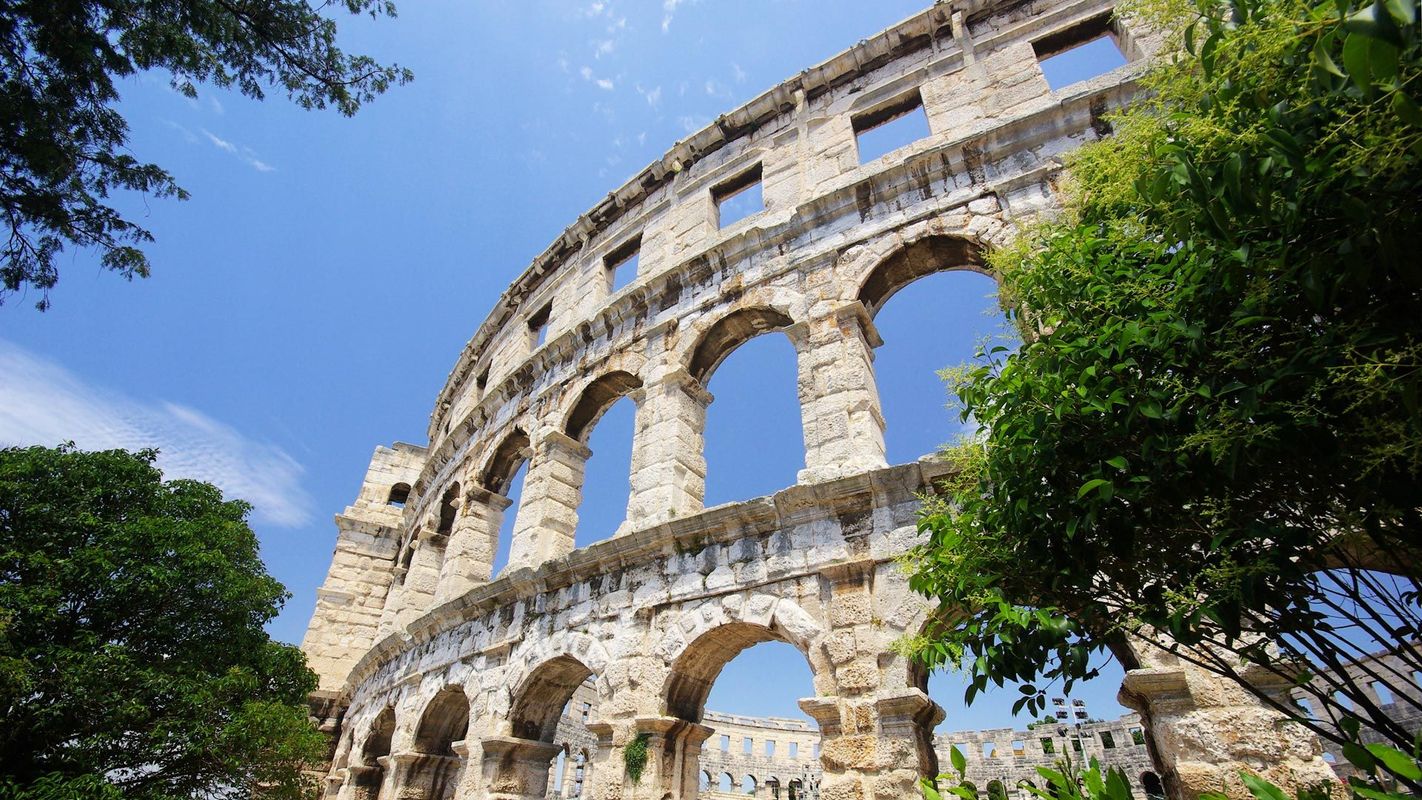 Nærbillede af den imponerende romerske arena i Pula, Kroatien, set nedefra med blå himmel i baggrunden. De velbevarede buer og stenfacader er omkranset af grønne træer, hvilket fremhæver kontrasten mellem antik arkitektur og frodig natur.