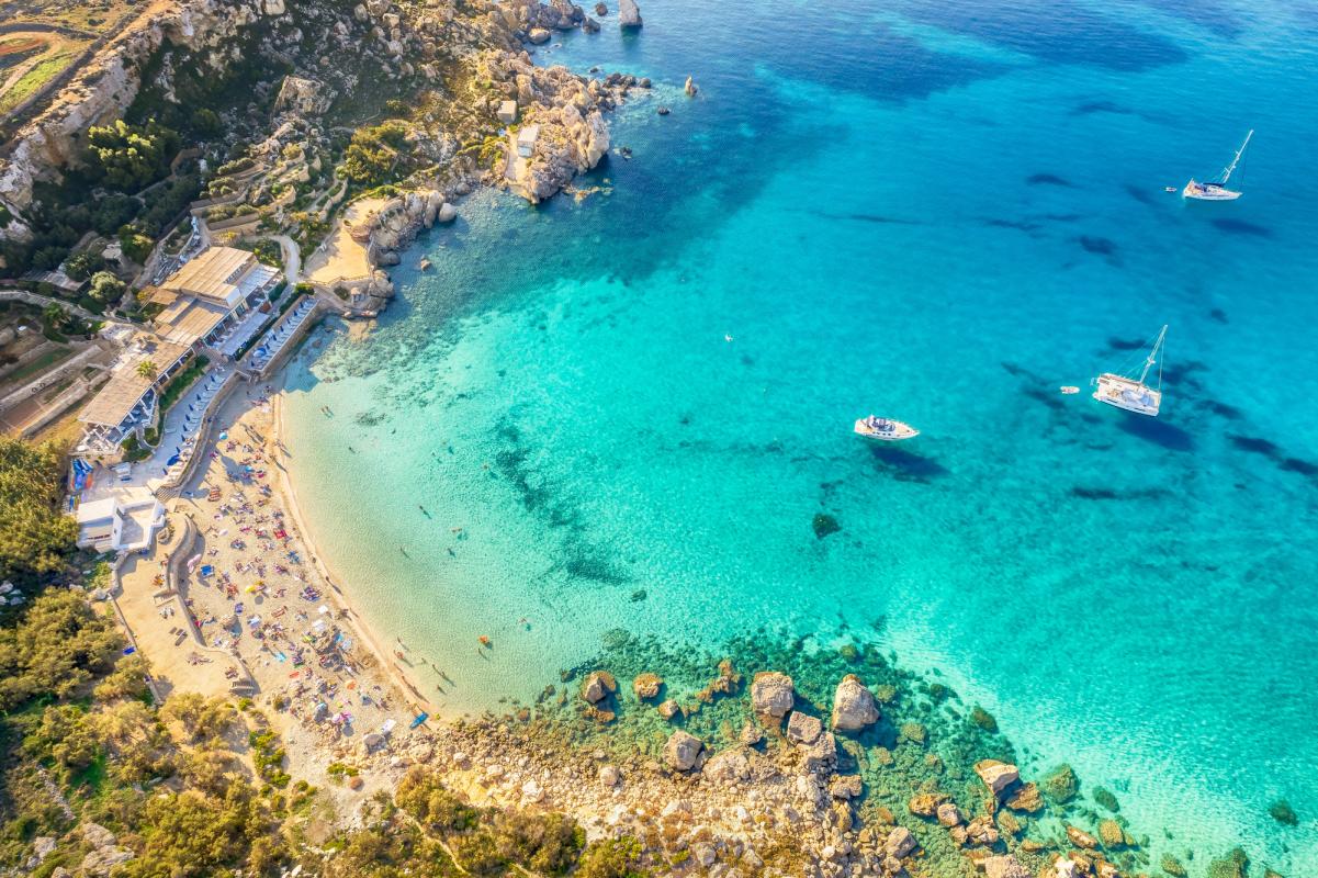 Klart turkosblått vatten med flera förankrade båtar i en lugn vik. På land syns en strand med färgglada parasoller och ett hotell med solstolar vid vattnet.