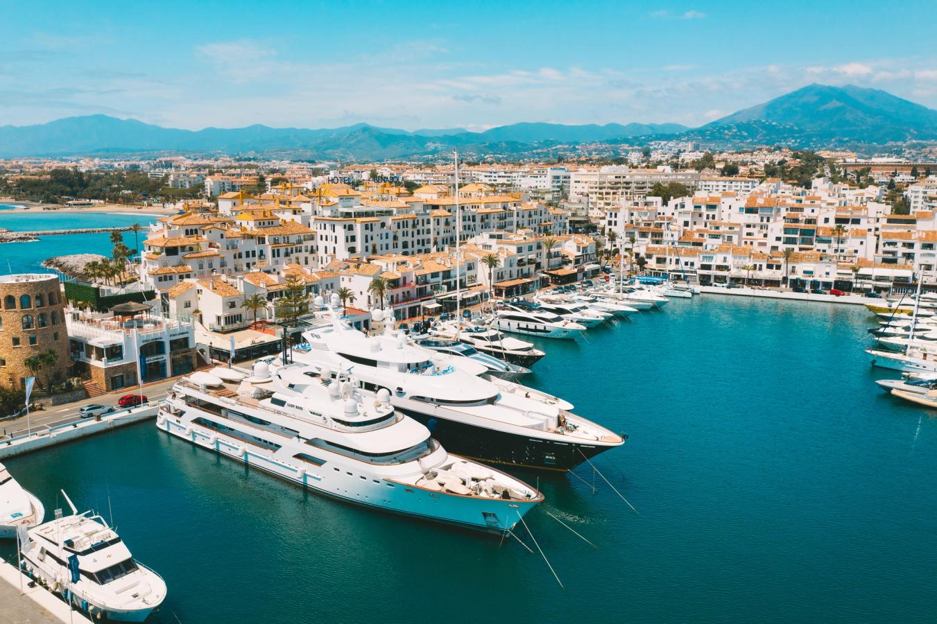 Puerto Banus marina hvor yachter og sejlbåde ligger side om side i det blå vand. I baggrunden ses byens hvidkalkede huse og i baggrunden rejser bjergene sig. 