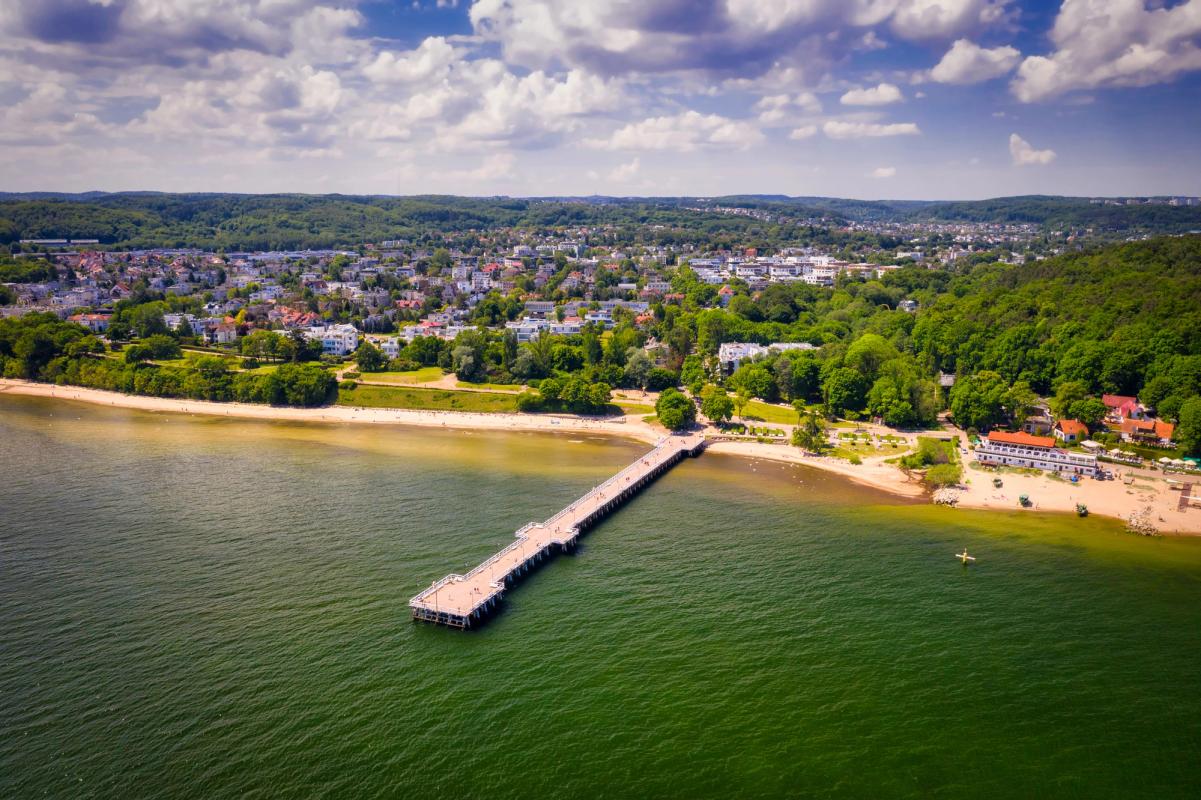 Bilden visar stranden Orłowo vid Gdynia, med en träbrygga en bra bit ut i havet. Havet är grönt med en kombination av skog och bebyggelse i bakgrunden av stranden.