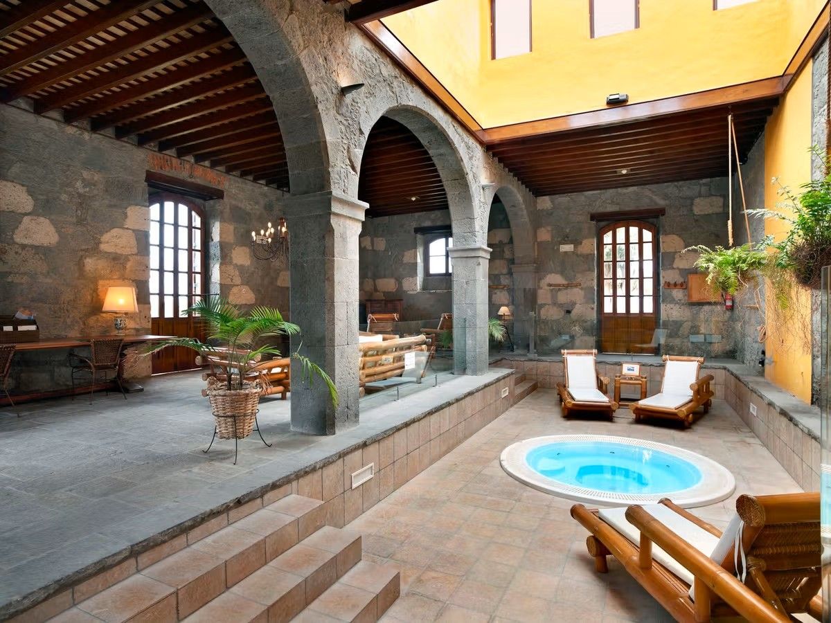 Et fællesareal på Hacienda del Buen Suceso. Her ser vi rødbrune gulvfliser med en lille jacuzzi i midten. Gulvet hæver sig til venstre med forskellige siddegrupper. Alt under buede bærende vægge og åben belysning fra loftet, omgivet af gule vægge.
