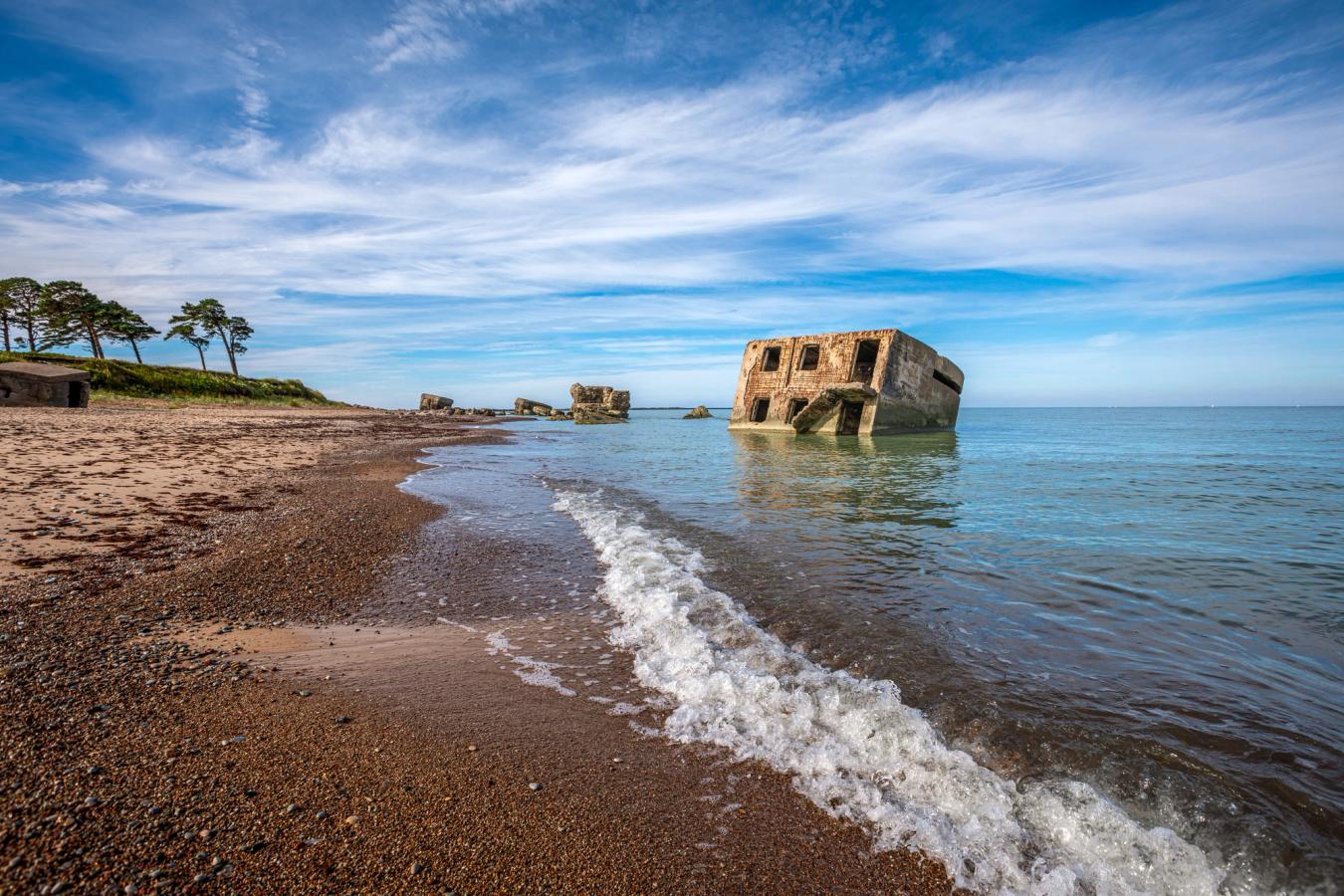 Bilden visar en strand vid Liepaja med små vågor från Östersjön och ett gammalt fort som ligger delvis ute i vattnet. I bakgrunden syns kusten med sand, enstaka träd och en klarblå himmel