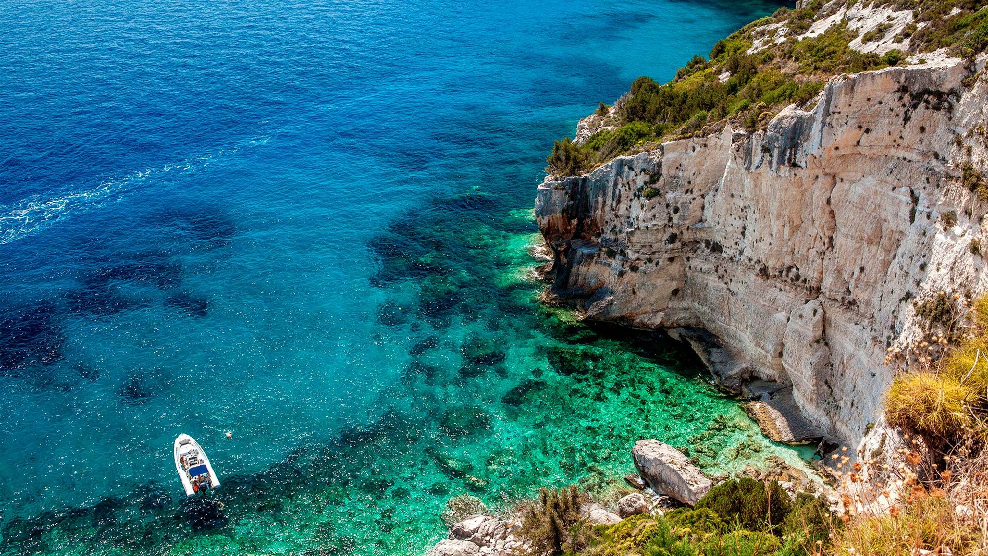 En liten motorbåt ligger for anker i klart turkisblått vann ved foten av en bratt, klippefylt kyst i Kroatia. Sollyset reflekteres i det gjennomsiktige havet, og grønn vegetasjon dekker toppen av klippene.