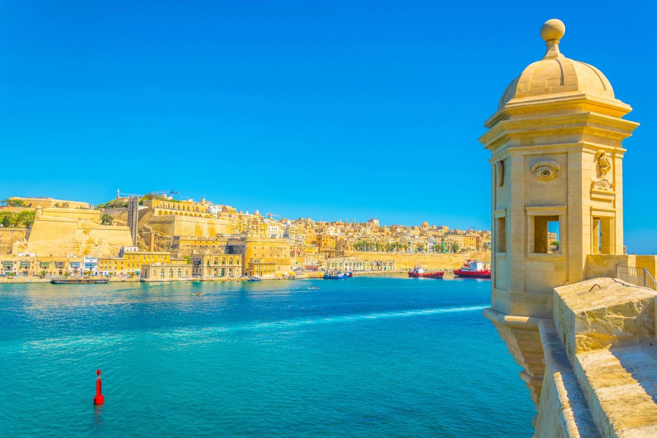 Bildet viser Upper Barakka gardens i Valletta på Malta. Med bybildet under en skyfri himmel til venstre, krystall blått vann i midten og med hjørne av eldre arkitektur til høyre.